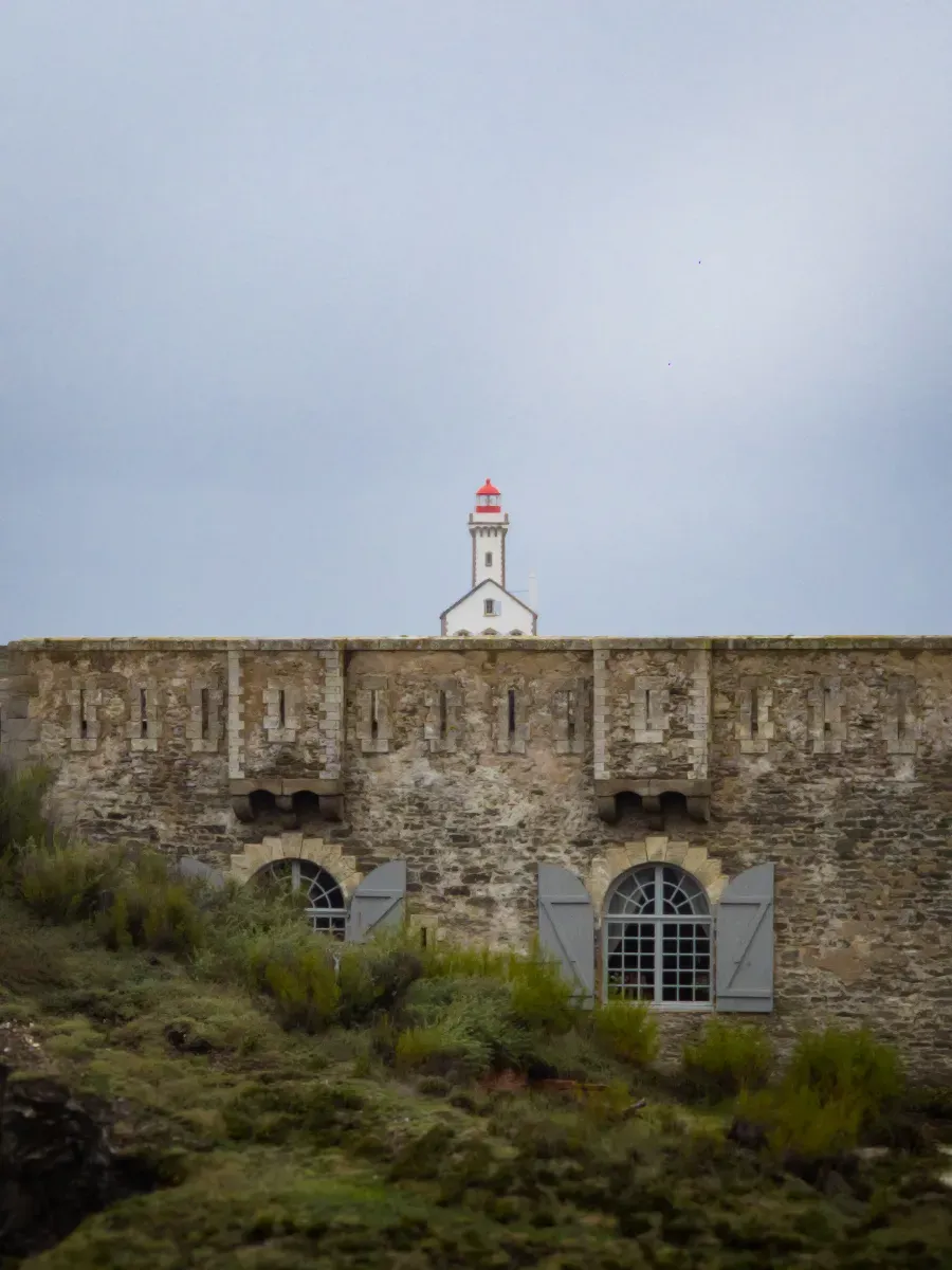 Phare blanc au toit rouge dépassant derrière le fort Sarah-Bernhardt de Belle-Île-en-Mer, sous un ciel couvert.