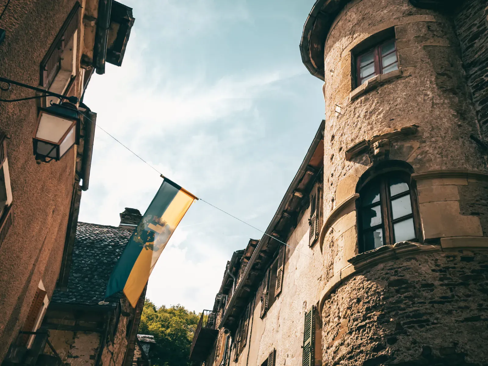 Vue en contre-plongée sur les façades en pierre d’Estaing avec un drapeau suspendu entre les bâtiments.
